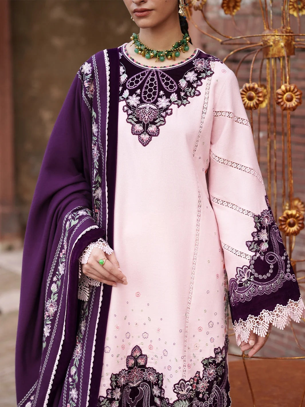 A woman models a light pink traditional South Asian outfit with a deep purple dupatta and ornate floral embroidery in purple, white, and green on the neckline and sleeves.