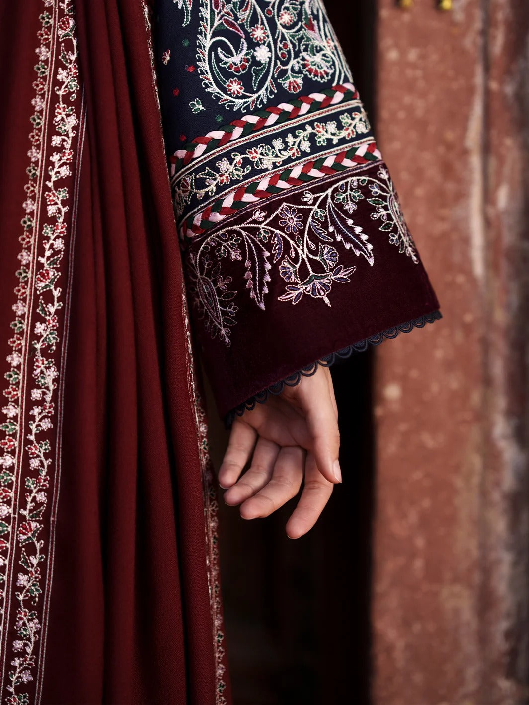 A close-up of a hand wearing a deep maroon embroidered sleeve with intricate floral embroidery and a scalloped edge, next to a matching maroon draped fabric.