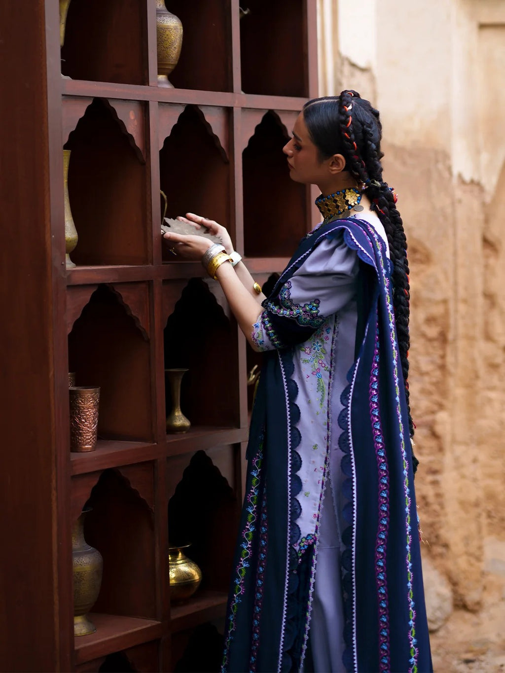A woman in traditional South Asian attire, featuring a navy blue embroidered dupatta and a light purple kurta, stands beside a dark wooden arched shelf in a rustic setting.