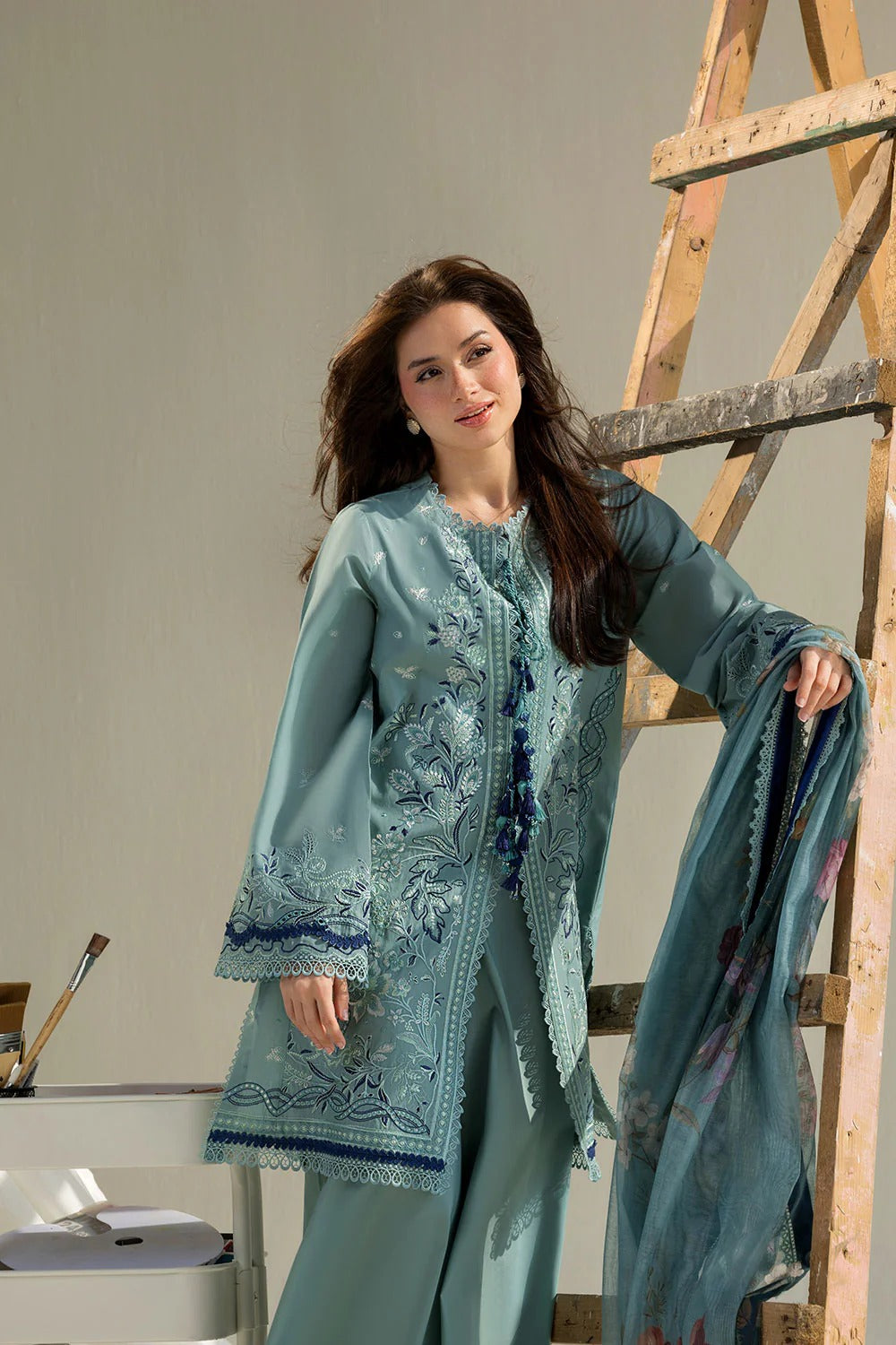 Woman wearing a light blue embroidered outfit standing next to a wooden ladder.