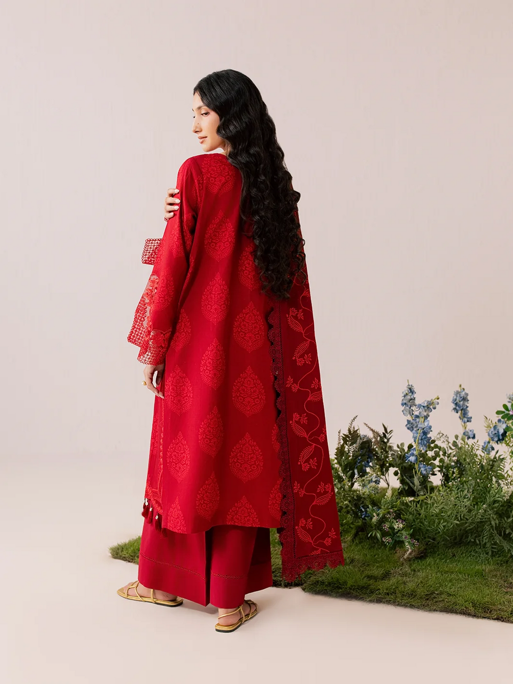 A woman wearing a red traditional outfit with matching pants and a long embroidered dupatta, photographed from the back against a plain beige studio background with a small decorative plant arrangement at the lower right.
