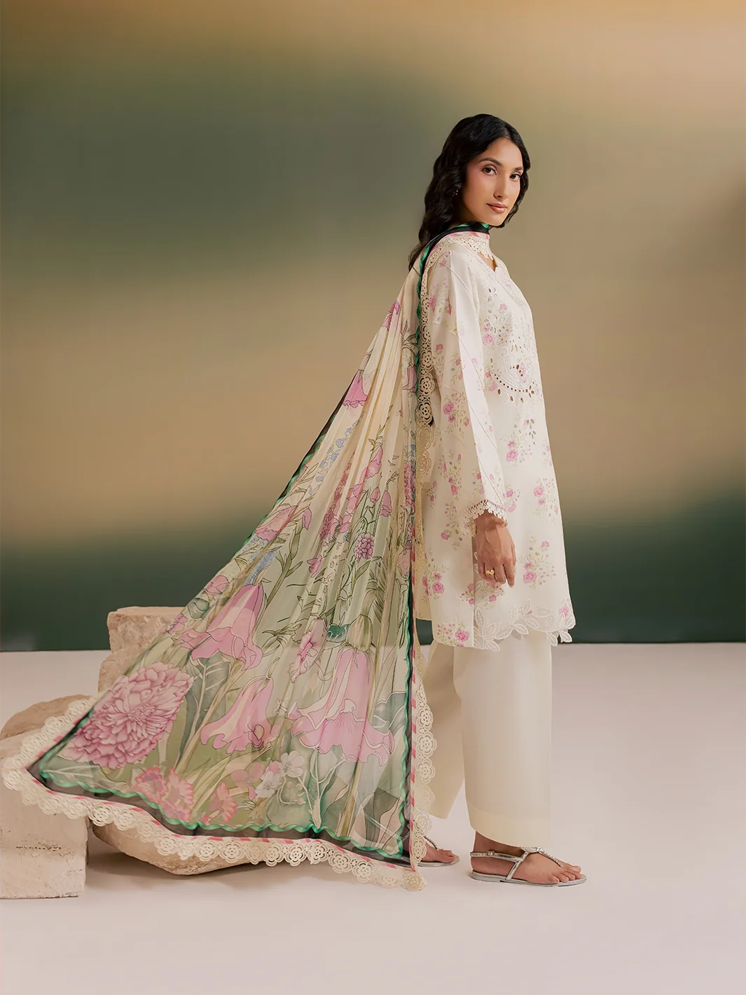 A woman models a traditional South Asian outfit consisting of an off-white embroidered kurta, matching wide-leg trousers, and a sheer floral dupatta with pink flowers and green leaves, set against a soft gradient studio background.