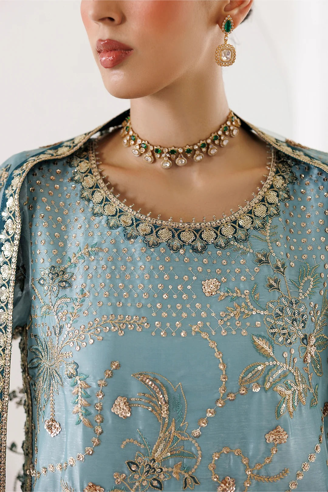 A close-up of a model wearing a light blue embroidered traditional outfit with intricate gold and silver beadwork, a matching embellished neckline, and a coordinating dupatta. The model is also wearing a gold and green beaded choker necklace and matching drop earrings.