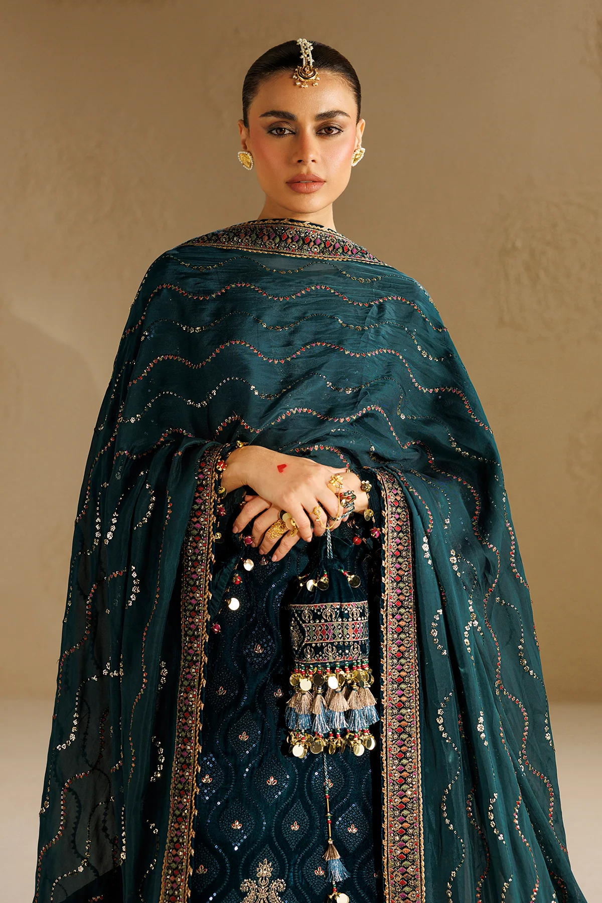 A woman models an ornate dark teal green traditional embroidered outfit with a matching sheer dupatta, decorated with intricate metallic embroidery, mirror work, and gold coin details against a warm beige background.