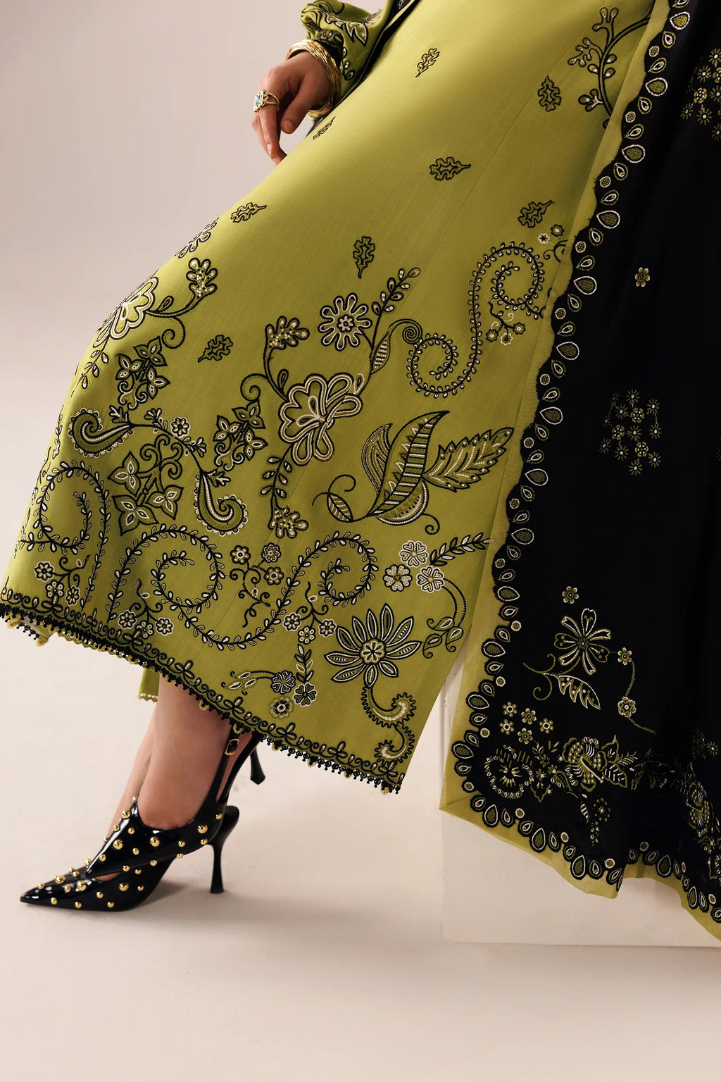 A close-up of a person wearing a bright yellow-green embroidered traditional outfit with a black dupatta and black studded high heels, photographed against a plain light studio background.