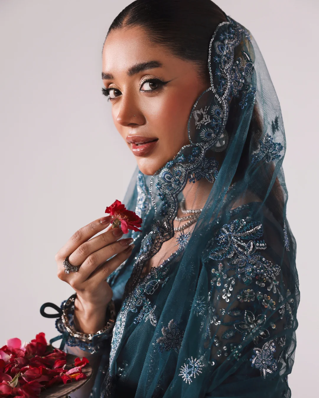 A woman wearing an ornate teal blue embroidered traditional outfit with a matching sheer veil, holding a red rose against a plain light background.