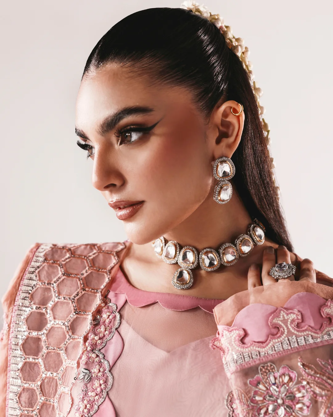 A close-up portrait of a woman wearing ornate silver-toned jewelry, including a choker necklace, matching drop earrings, and rings, paired with a pink embroidered traditional outfit against a light neutral background.