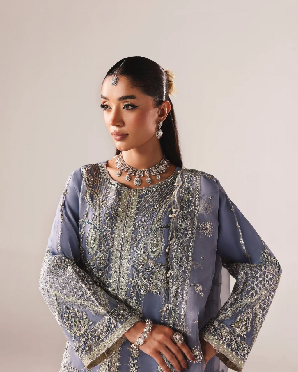 A woman models a heavily embellished traditional outfit in a studio setting. The garment is a muted dusty blue with intricate silver and gold embroidery, beadwork, and mirror work across the sleeves and front panel.