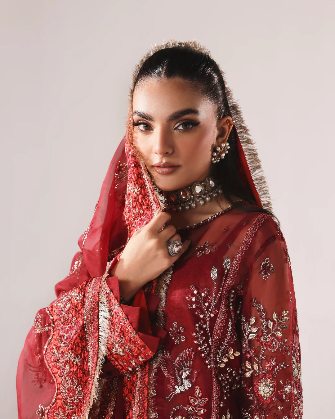 A woman wearing an ornate red embroidered traditional outfit with a matching sheer dupatta, embellished with intricate metallic floral embroidery and fringe details, set against a plain light gray studio background.