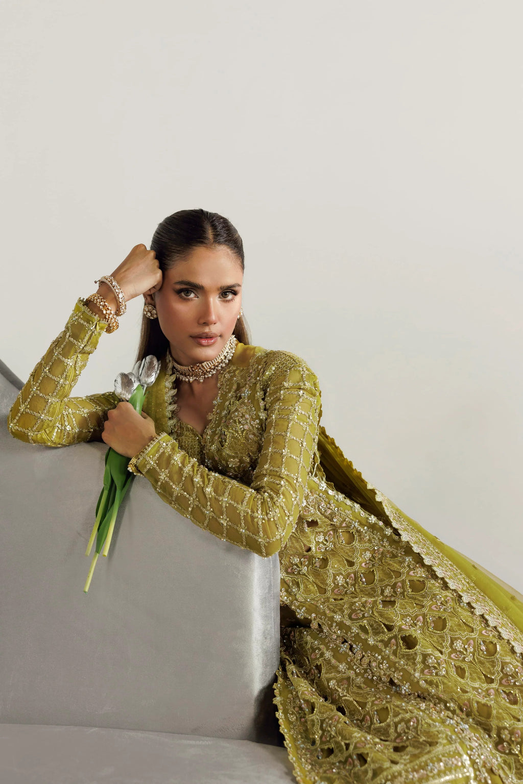 A woman models a traditional olive green embroidered kurta set with a matching dupatta, leaning against a light gray sofa against a plain off-white wall.