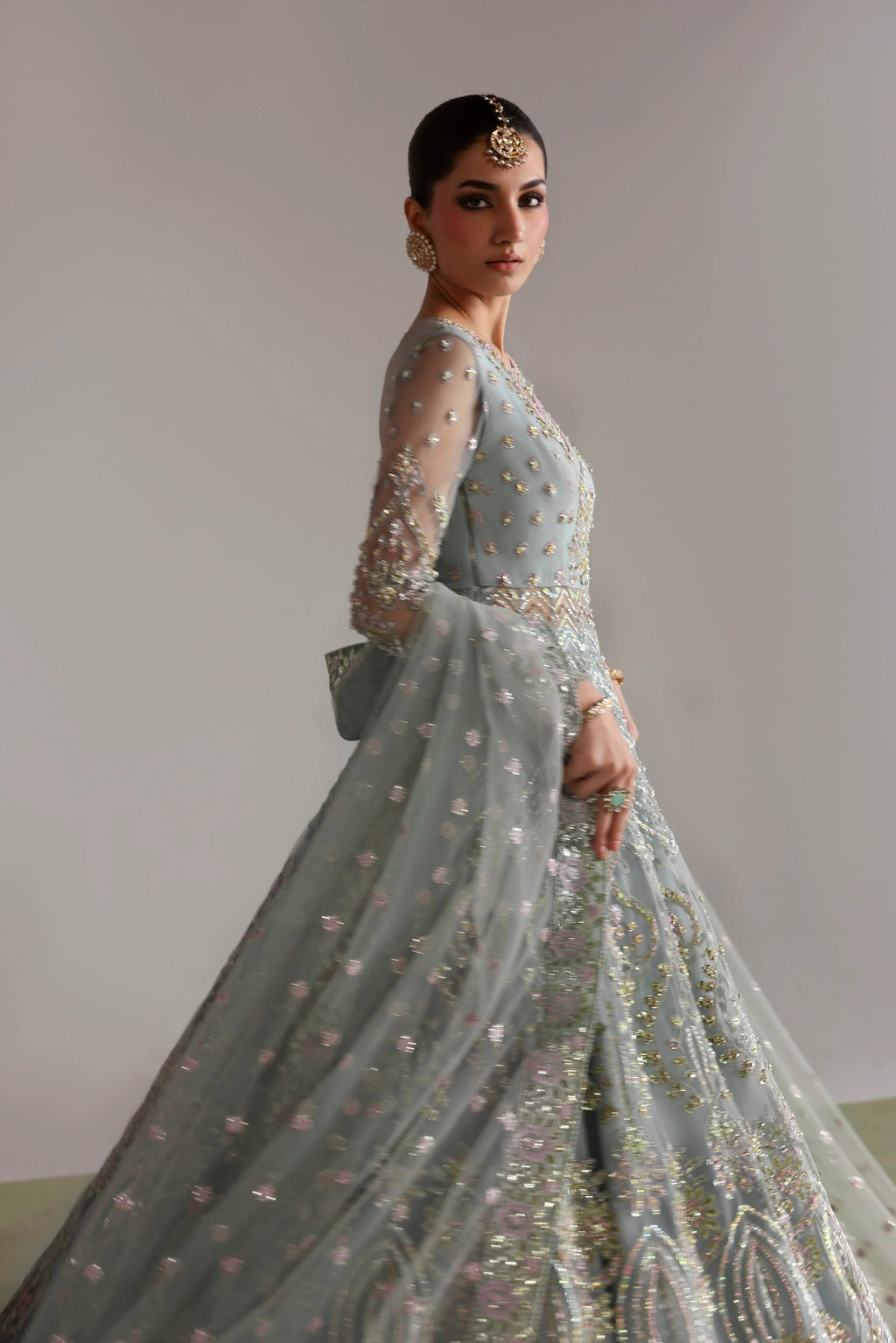 A woman modeling a light blue-green embellished traditional gown with sheer long sleeves, intricate beadwork, and a full flared skirt, photographed against a plain light gray wall.