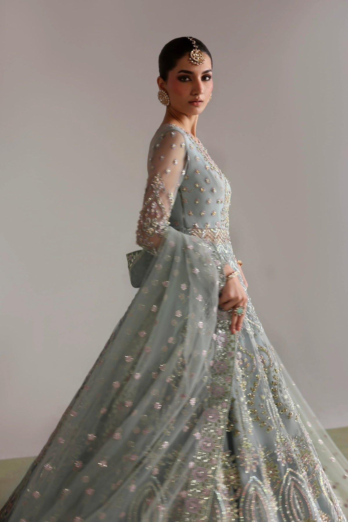 A woman modeling a light blue-green embellished traditional gown with sheer long sleeves, intricate beadwork, and a full flared skirt, photographed against a plain light gray wall.