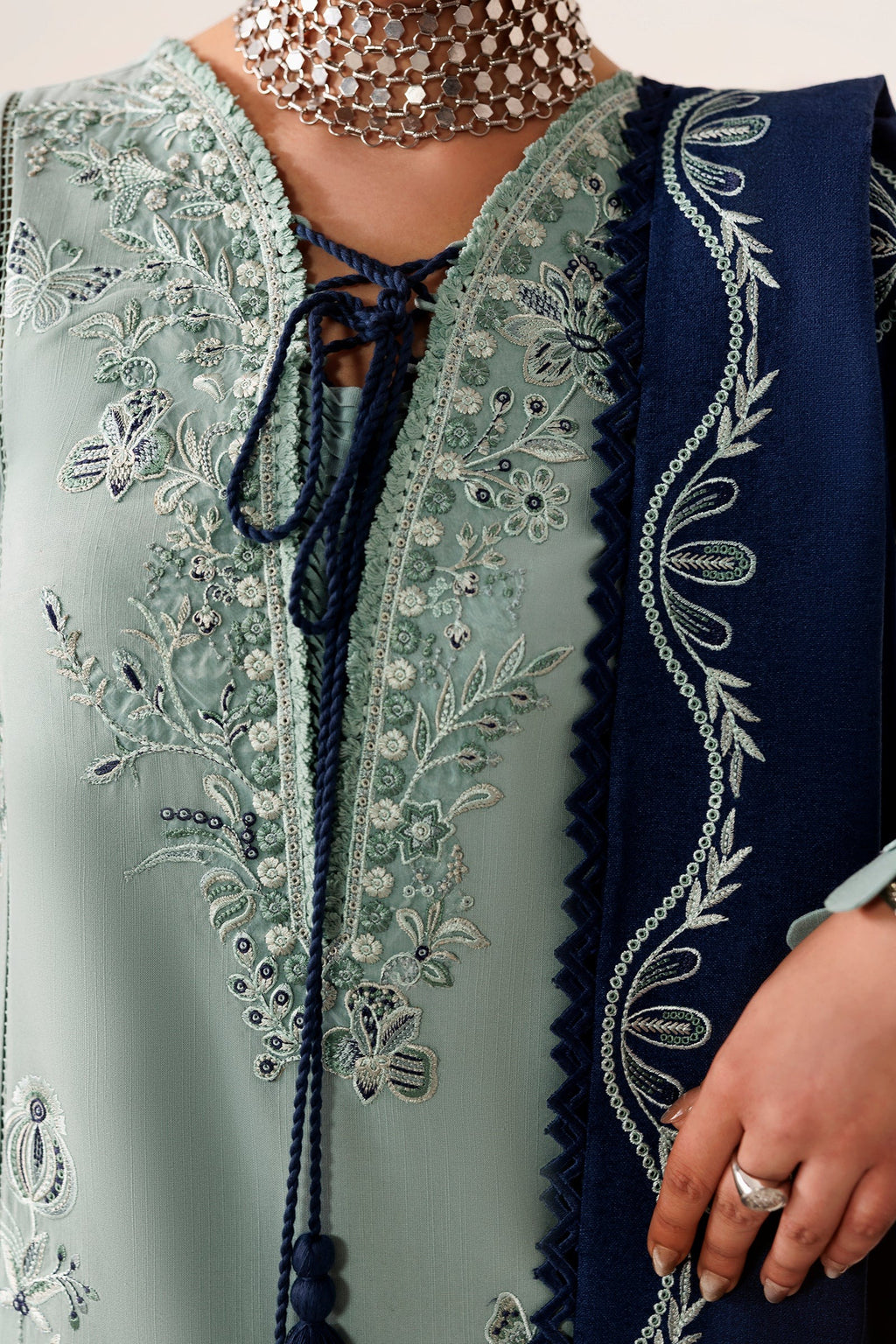 A close-up of a traditional embroidered outfit featuring a sage green kurta with intricate silver and white floral embroidery, a navy blue tie at the neckline, and a matching navy blue dupatta draped over the shoulder.