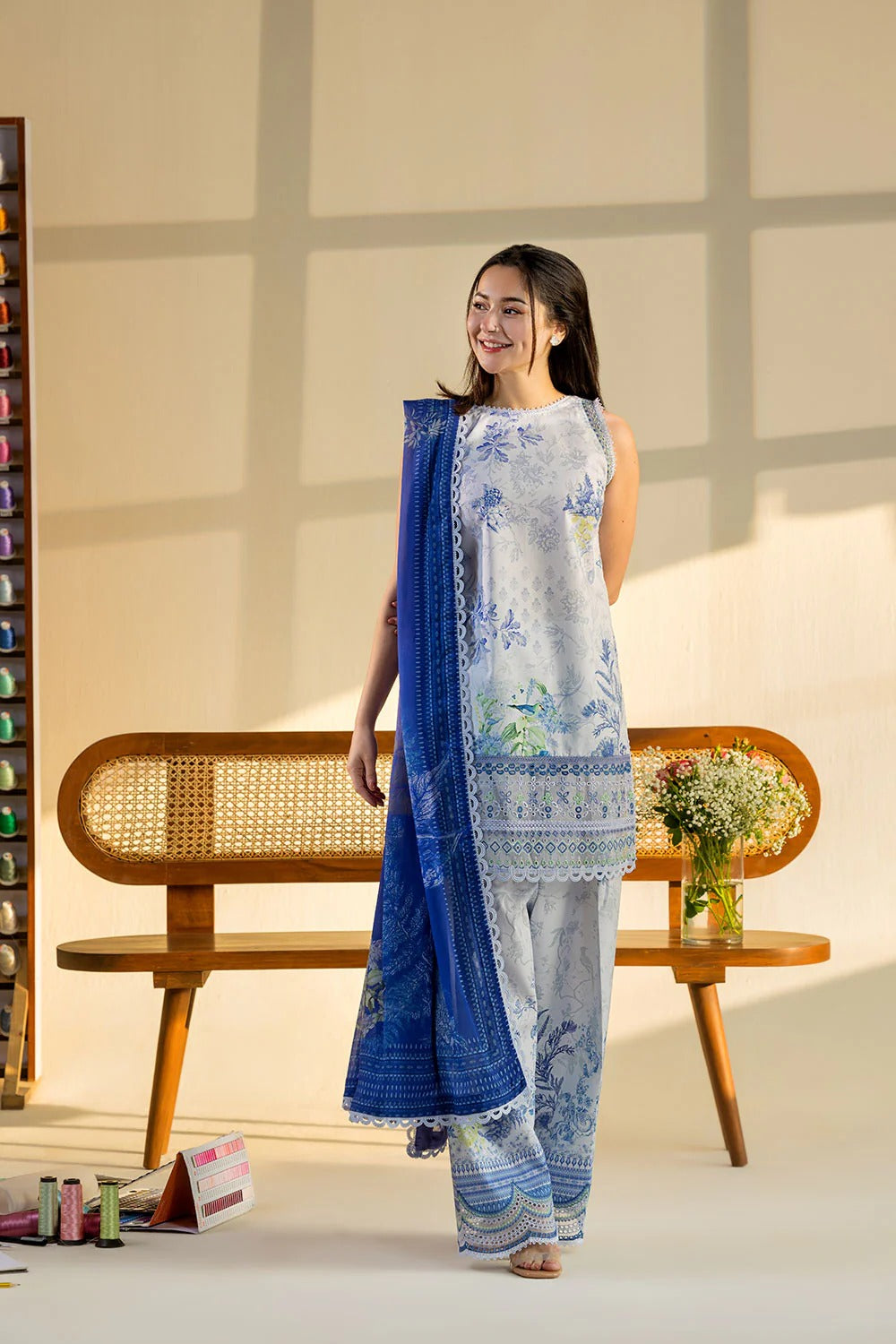 Woman in a blue and white traditional outfit standing in a room with a wooden bench and decorative items.