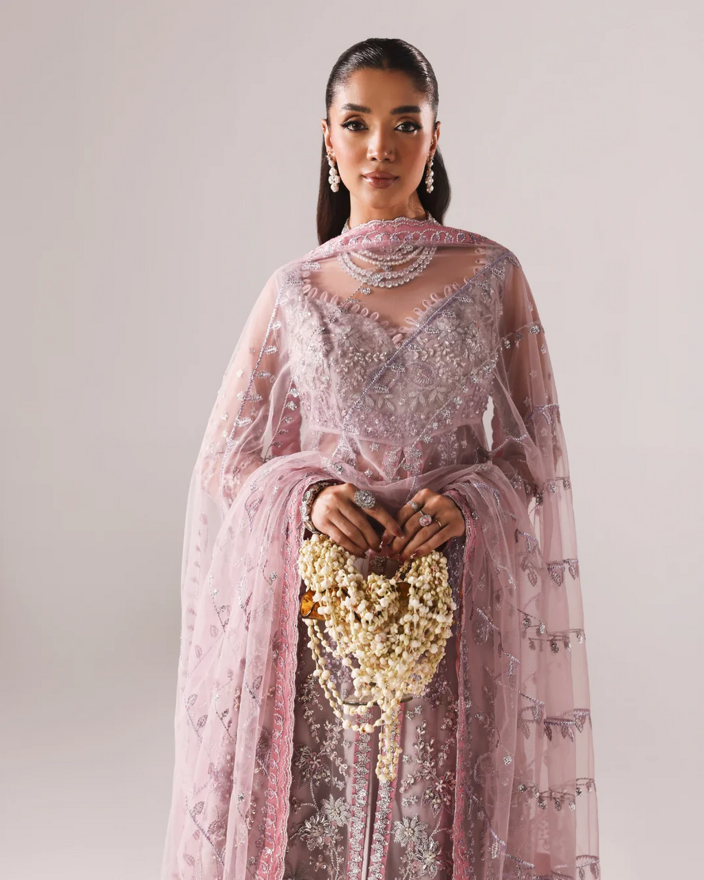 A woman modeling a heavily embellished light pink traditional outfit with silver embroidery, a matching sheer dupatta, layered pearl necklaces, and a pearl waist garland against a plain light gray studio background.