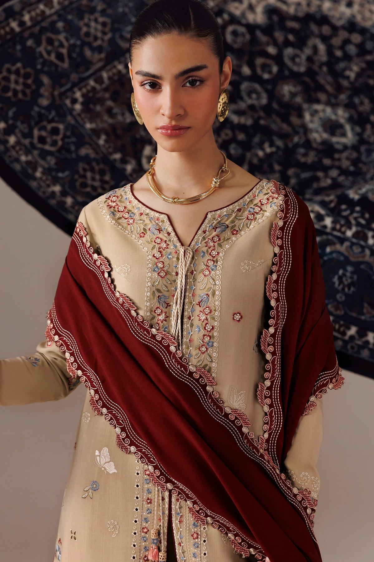 A woman models a traditional South Asian salwar kameez suit set. The outfit features a beige embroidered kameez top with floral patterns, paired with a deep maroon dupatta that has a decorative embroidered border.