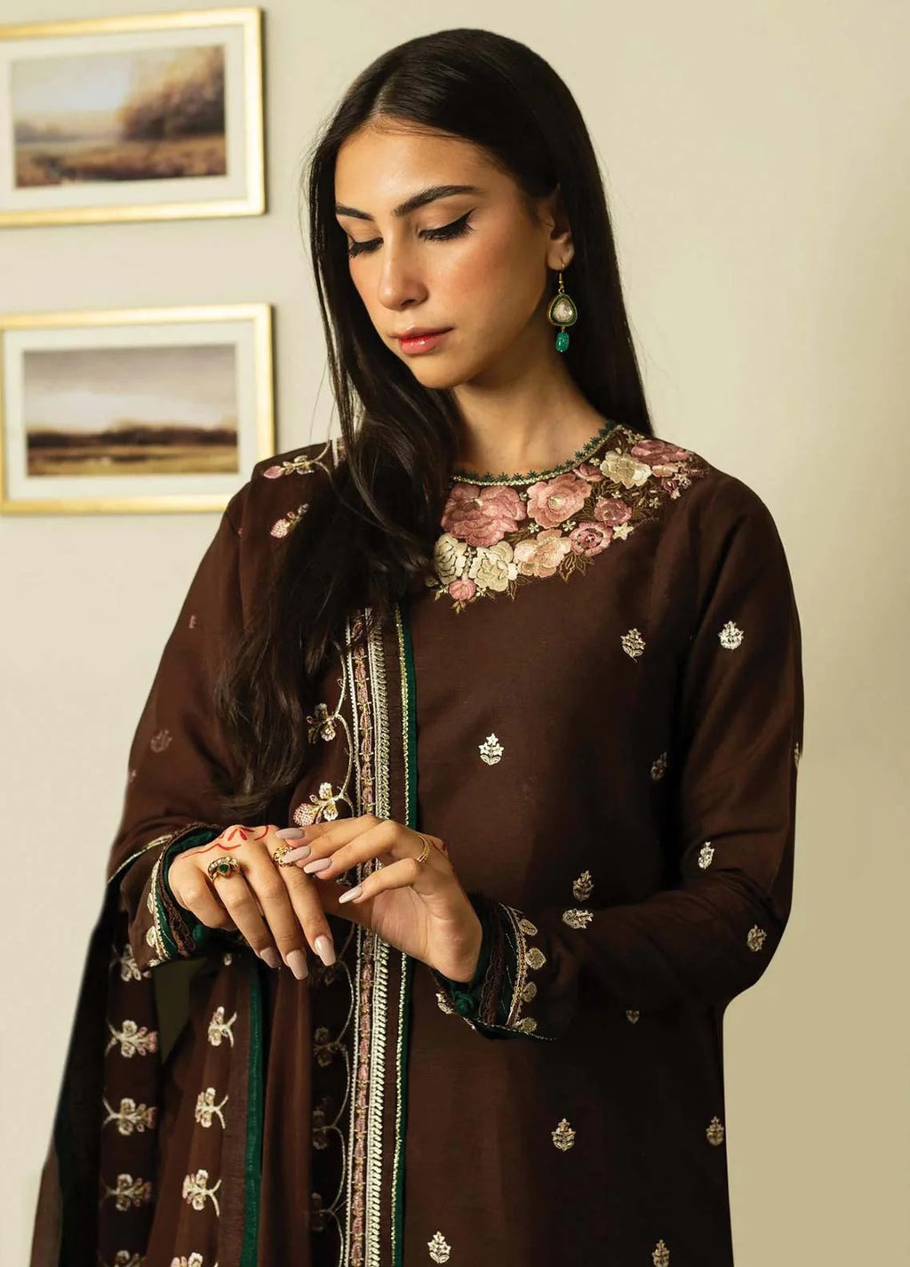 A woman wearing a dark brown traditional embroidered outfit with floral embroidery around the neckline, metallic motifs on the sleeves, and a matching dupatta, posed against a light beige wall with framed pictures.