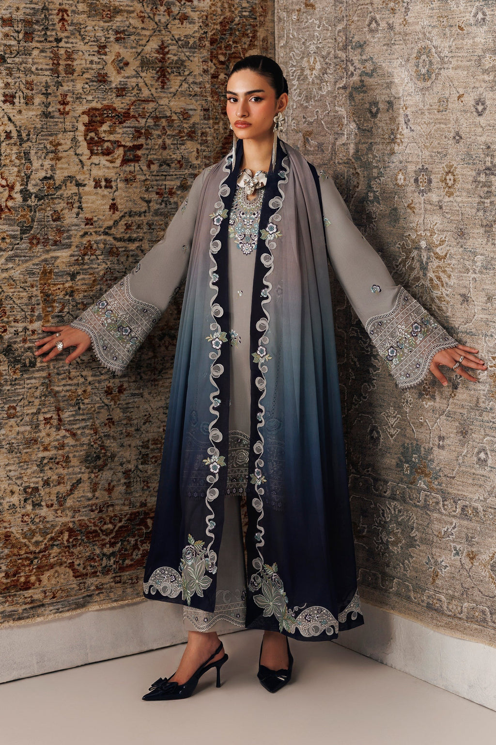 A woman models a traditional long gray and navy blue ombre embroidered dress with a matching sheer scarf, standing against a patterned rug.