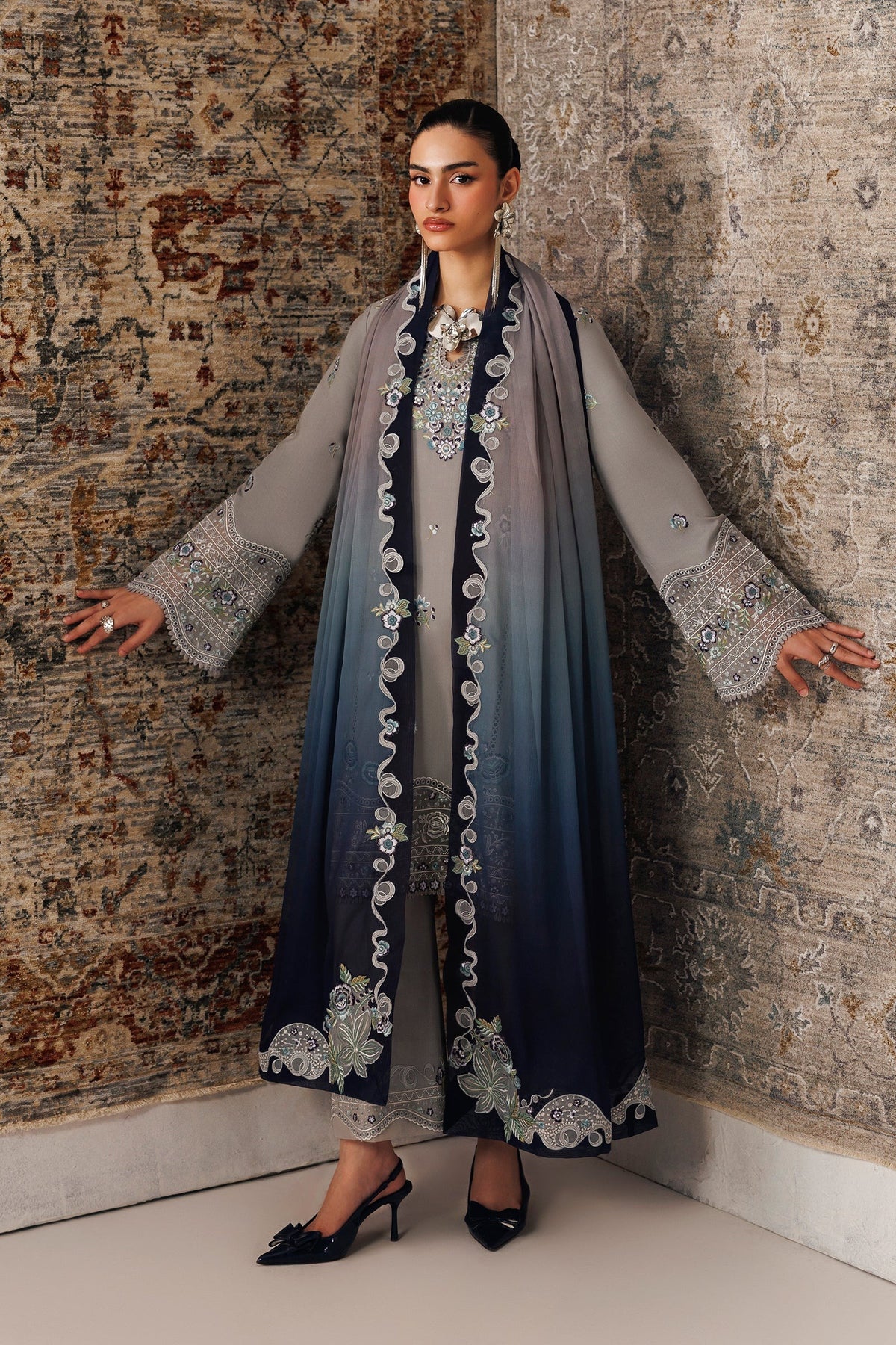 A woman models a traditional long gray and navy blue ombre embroidered dress with a matching sheer scarf, standing against a patterned rug.