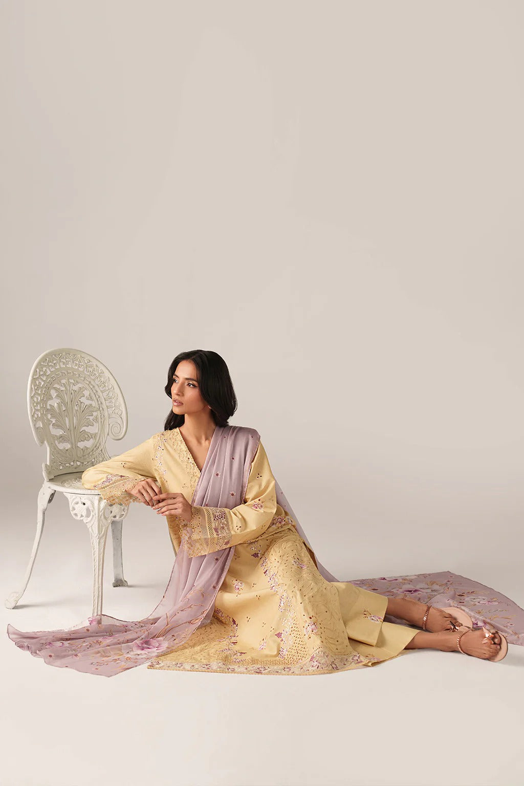 A woman models a traditional three-piece ethnic outfit consisting of a light yellow kurta, a lavender-pink dupatta, and matching trousers, posed next to a white decorative chair against a solid light beige background.