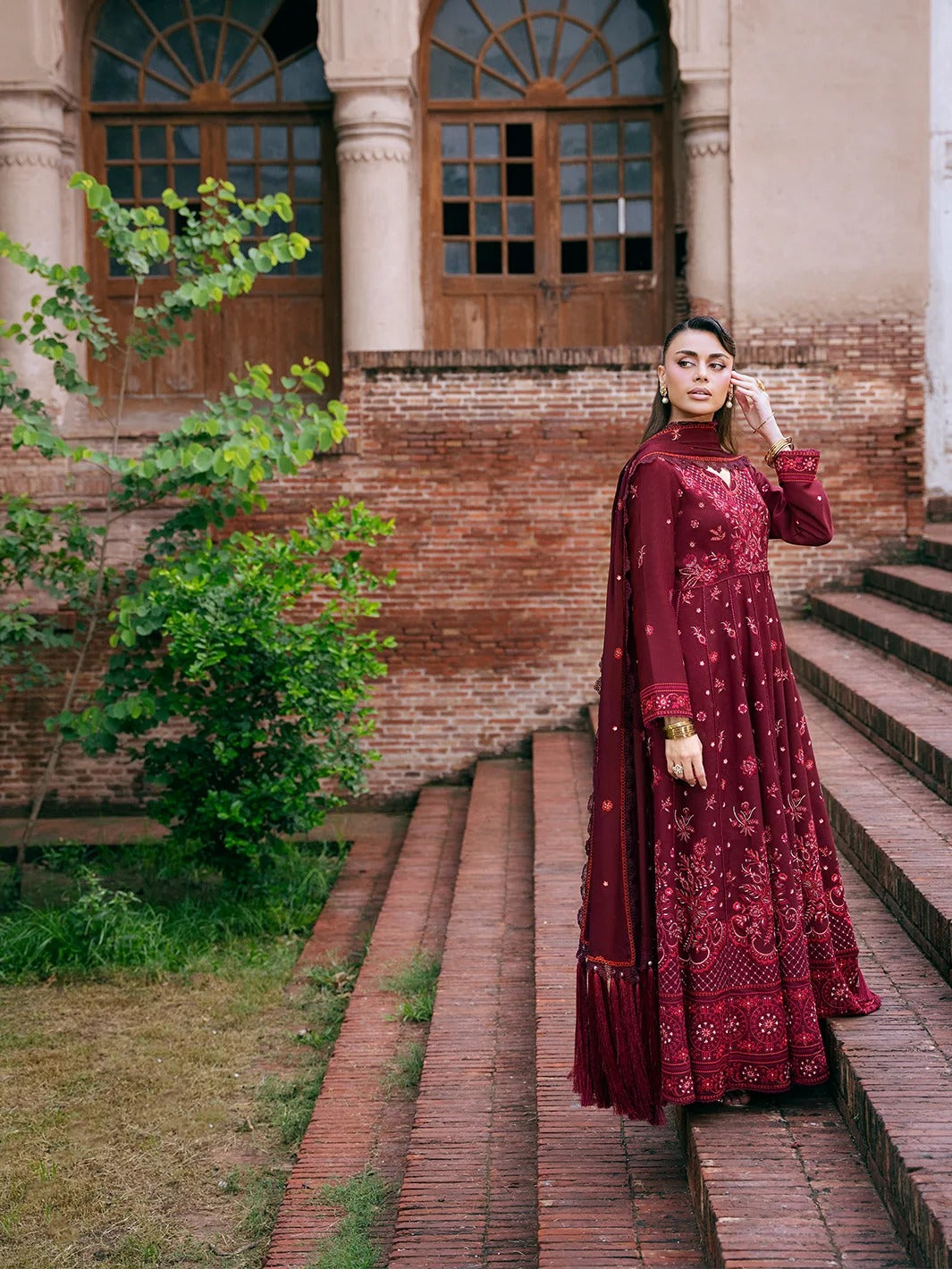A woman wearing a long maroon embroidered traditional dress stands on outdoor brick steps in front of a building with arched windows and columns.