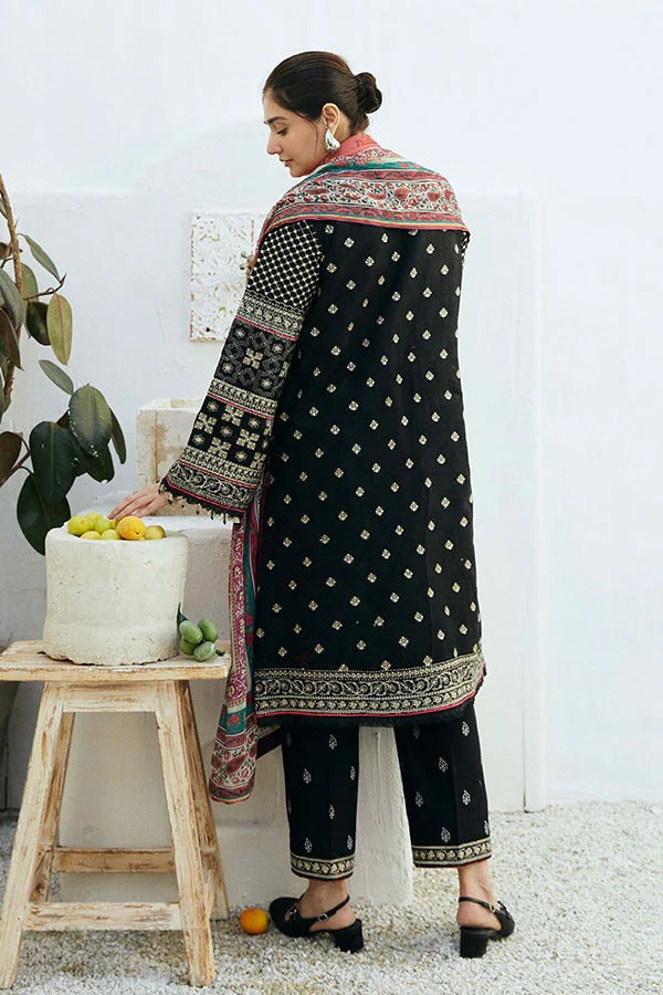 A woman modeling a black three-piece salwar kameez suit with intricate multicolor borders and white printed motifs, standing next to a small wooden stool with a white pot of lemons.