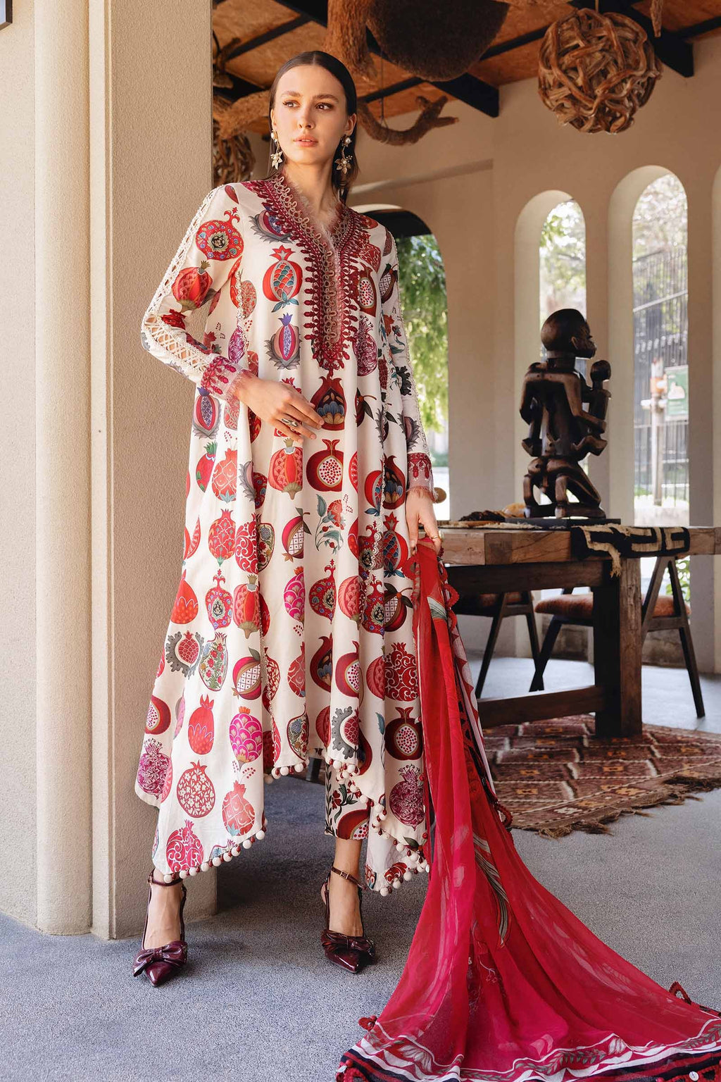 Woman in a patterned dress with a red scarf standing in a room with a table and statue.