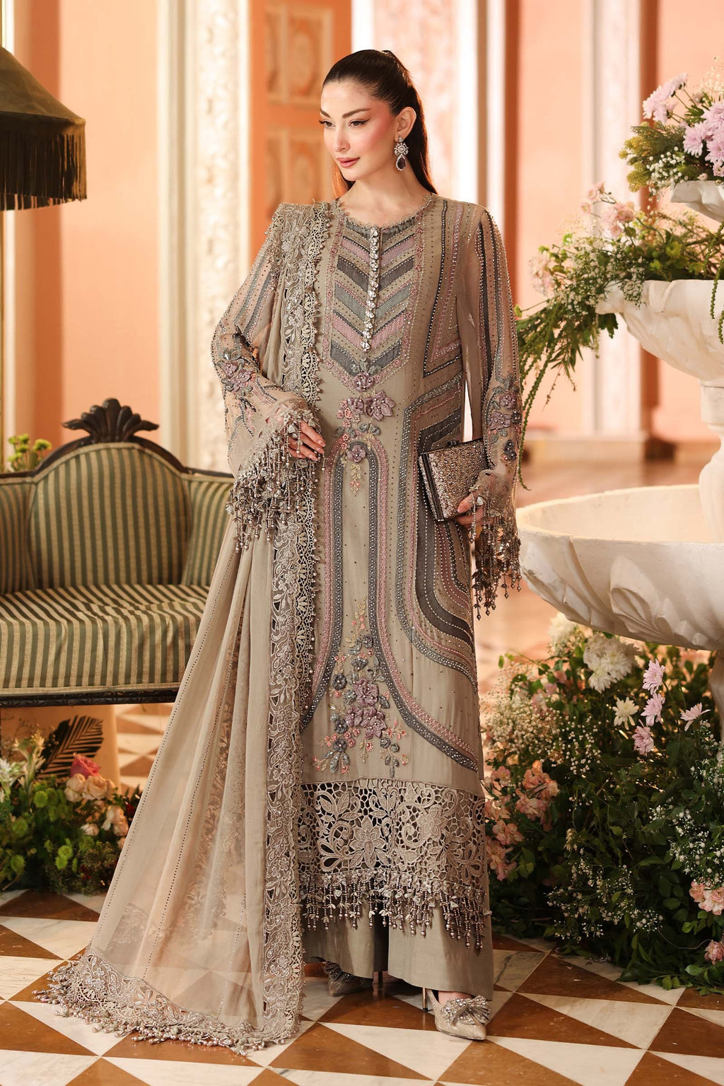 A woman models a traditional three-piece salwar kameez suit in a warm beige/taupe color with intricate silver and gold embroidery, set against an elegant indoor backdrop with a sofa and floral arrangements.