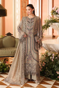 A woman models a traditional three-piece salwar kameez suit in a warm beige/taupe color with intricate silver and gold embroidery, set against an elegant indoor backdrop with a sofa and floral arrangements.