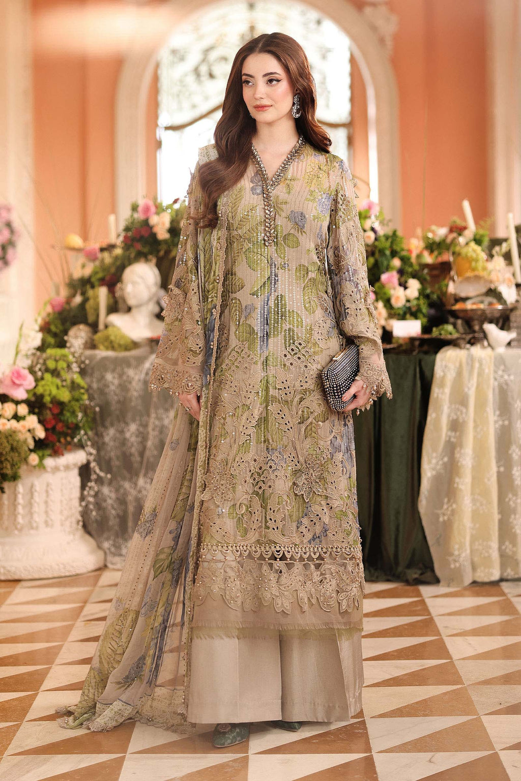 A woman models a traditional three-piece ethnic outfit with a beige base and green floral embroidery, featuring a long kurta, wide-leg trousers, and a matching dupatta, set against an elegant indoor backdrop with floral arrangements.