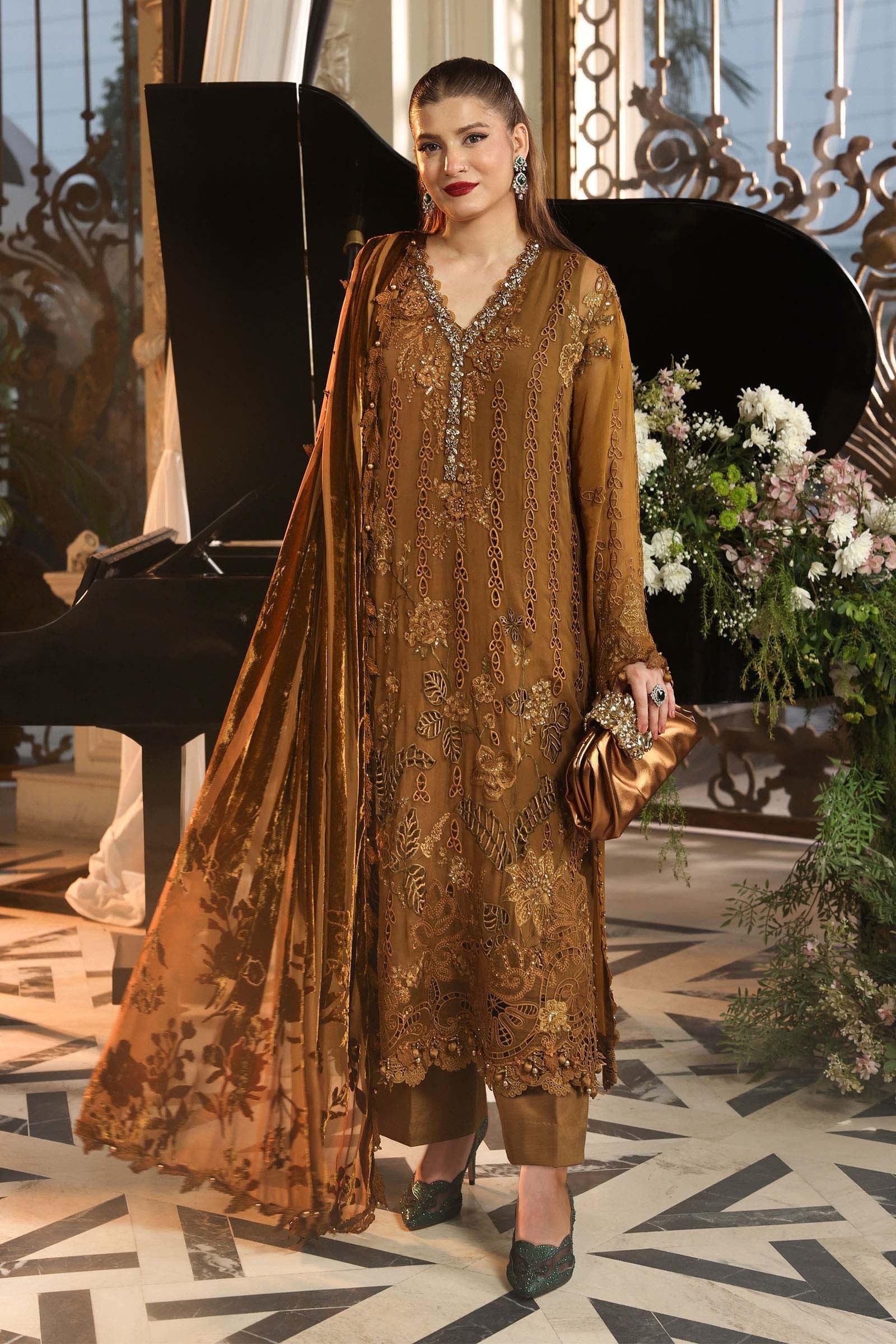 A woman models a traditional brown embroidered salwar kameez suit with a matching dupatta, standing in an elegant indoor setting with a black grand piano and floral arrangements in the background.