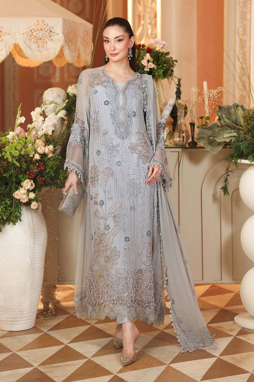 A woman models a light blue-gray traditional embroidered dress with a matching sheer dupatta and clutch, set in an elegant indoor room with floral arrangements.