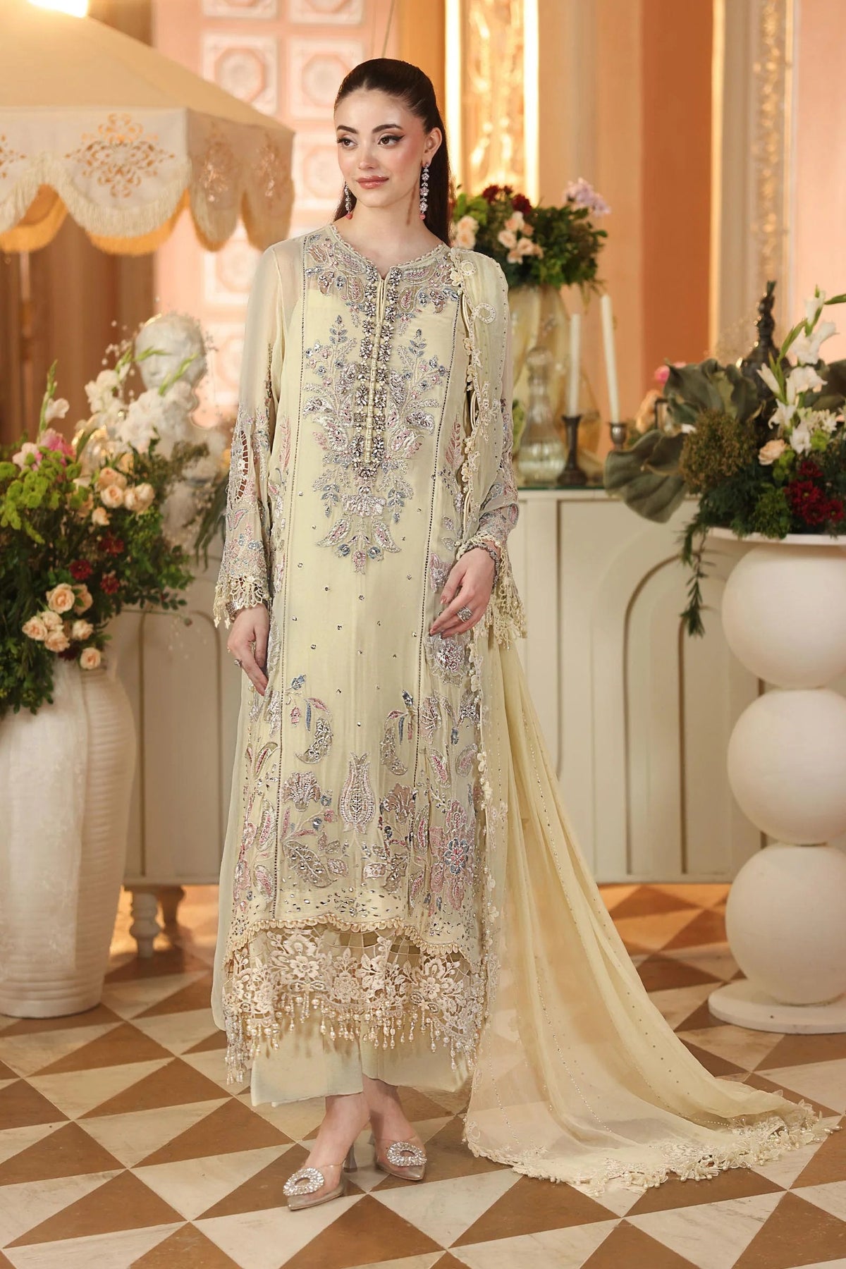 A woman models a traditional South Asian salwar kameez suit in a warm beige/cream color with intricate silver and blue embroidery. The outfit includes a long kurta, matching trousers, and a sheer dupatta, set against an elegant indoor background with floral arrangements.