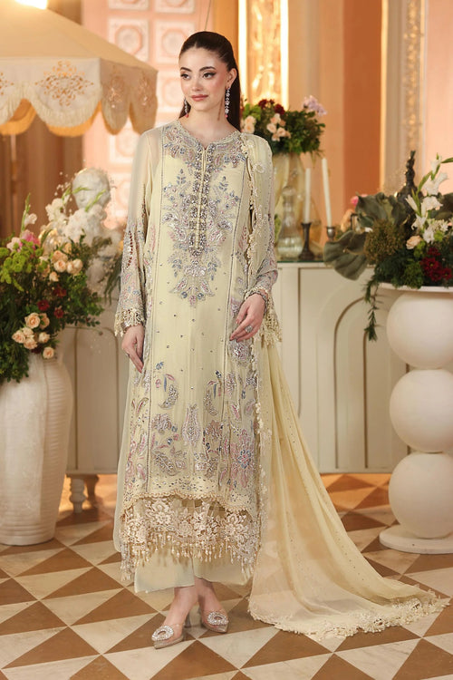 A woman models a traditional South Asian salwar kameez suit in a warm beige/cream color with intricate silver and blue embroidery. The outfit includes a long kurta, matching trousers, and a sheer dupatta, set against an elegant indoor background with floral arrangements.