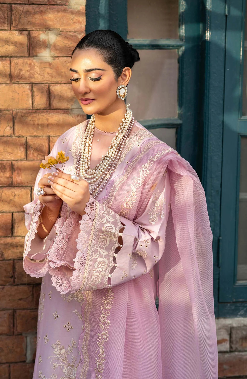 A woman wearing a light pink traditional outfit with intricate gold embroidery and layered pearl necklaces, standing against a brick wall and dark teal window frame.