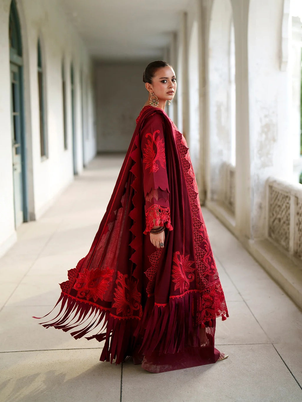 A woman wearing a deep red embroidered traditional dress with a matching sheer cape and fringe details, standing in a sunlit outdoor corridor with white columns.