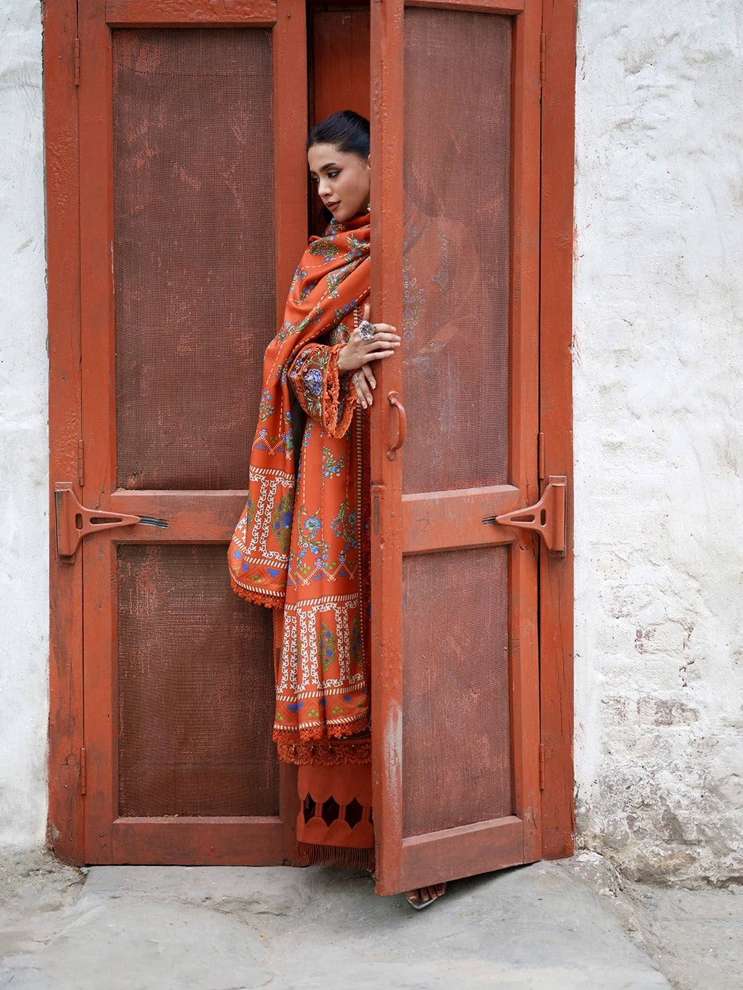 A woman wearing a vibrant orange and red patterned shawl and matching dress stands partially hidden behind an open reddish-brown wooden door against a white textured wall.