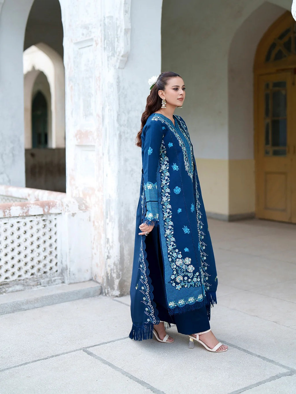 A woman models a traditional navy blue salwar kameez suit with intricate white and light blue floral embroidery, standing in an outdoor architectural setting with arches.