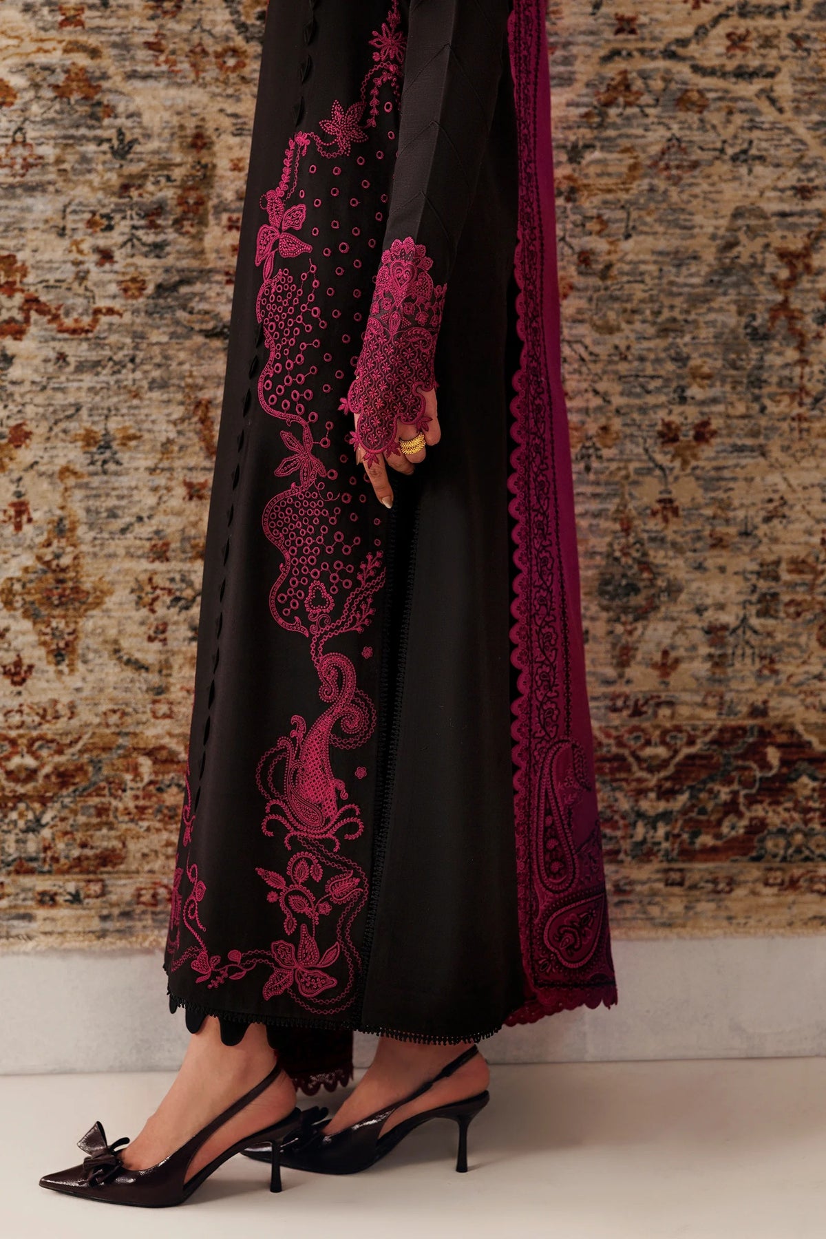 A person wearing a black traditional outfit with bright magenta embroidery and a matching embroidered dupatta, paired with black slingback heels, standing against a patterned rug.