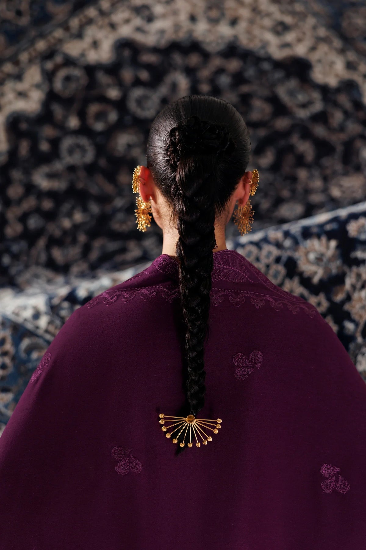 A woman viewed from behind wearing a deep purple embroidered garment with a long braided ponytail and a gold fan-shaped hair ornament, set against a dark patterned rug.