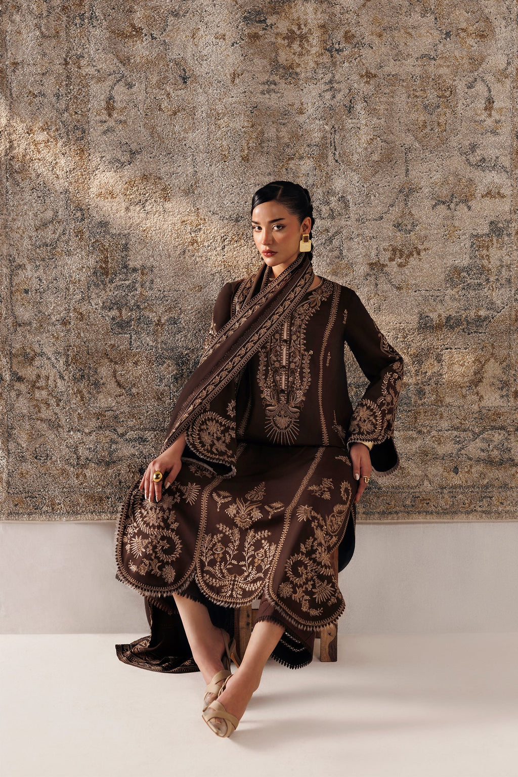 A woman models a dark brown traditional outfit with intricate gold embroidery, seated against a textured beige and brown wall backdrop.