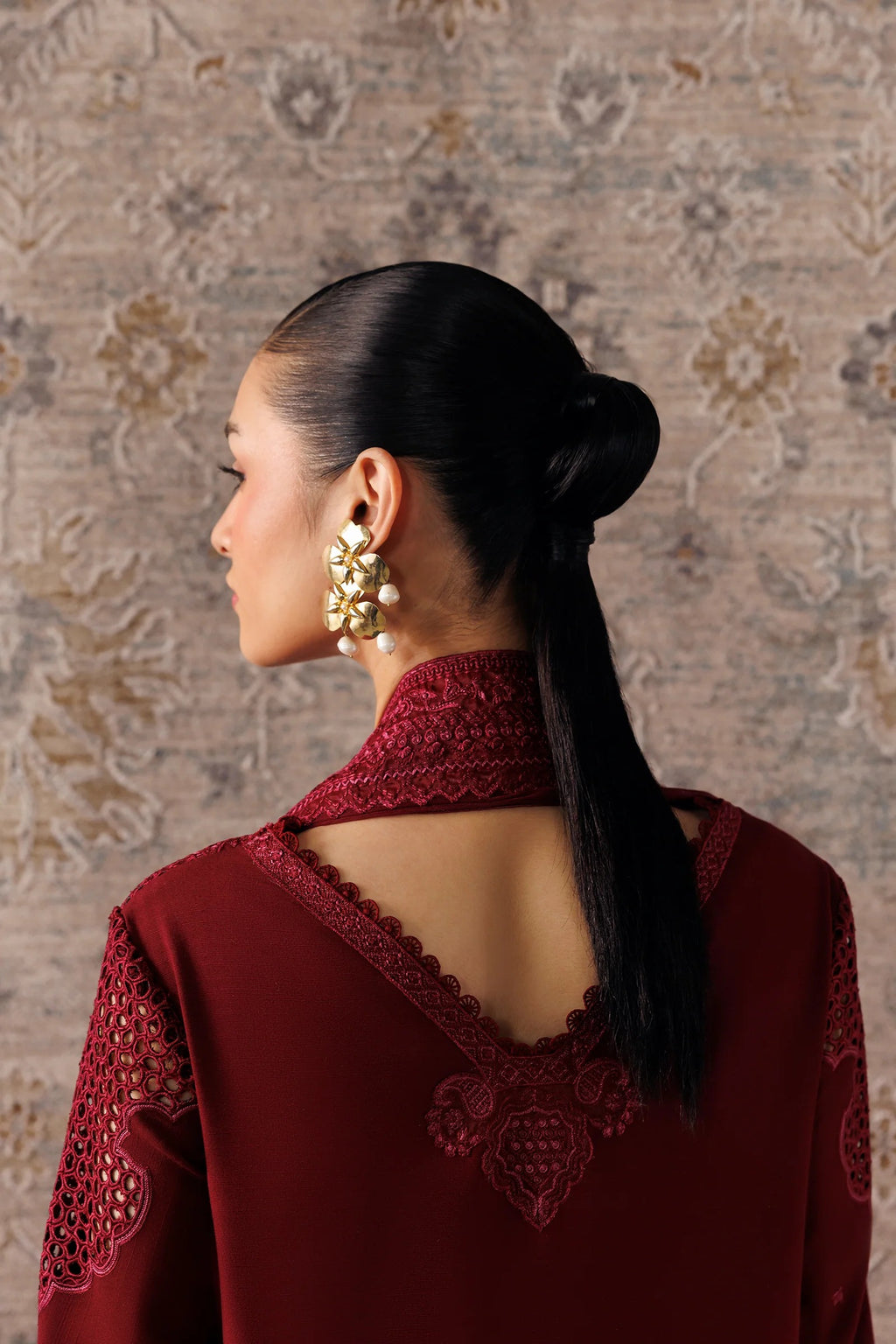 A woman shown from the back wearing a deep maroon embroidered top with a high lace-up collar and a long low ponytail, paired with large gold statement earrings, set against a patterned beige wall.