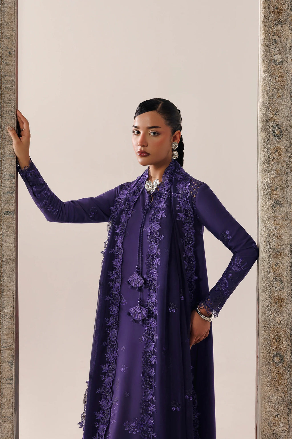 A model wearing a deep purple traditional outfit with matching embroidered outer layer, standing in front of a neutral beige background framed by stone pillars.