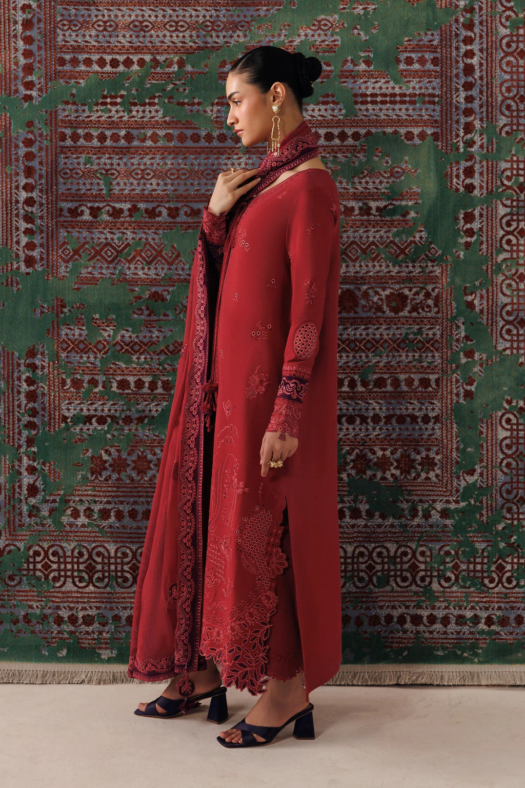 A woman models a traditional red Indian outfit with a matching dupatta, featuring intricate embroidery and a side slit, posed in front of a patterned green and red rug.