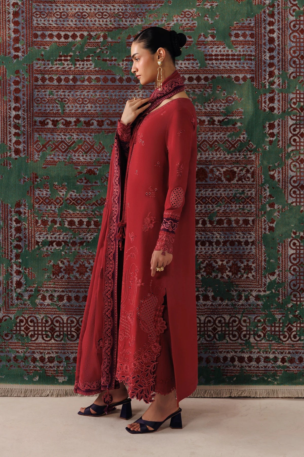 A woman models a traditional red Indian outfit with a matching dupatta, featuring intricate embroidery and a side slit, posed in front of a patterned green and red rug.