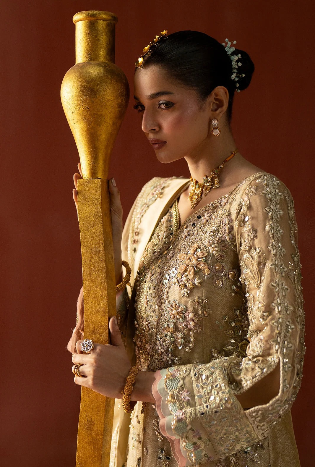 A woman in an ornate beige embellished traditional outfit holds a large gold vase against a warm reddish-brown studio background.