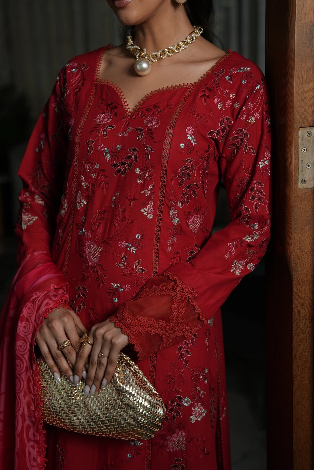 Woman in a red embroidered dress holding a gold clutch, wearing a pearl necklace.