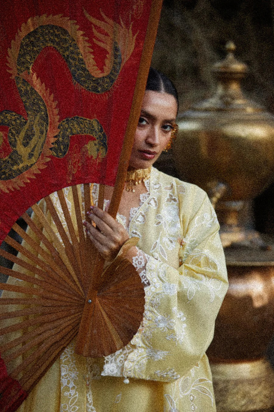 Woman in traditional attire holding a fan with a dragon design, standing in front of a large metallic pot.
