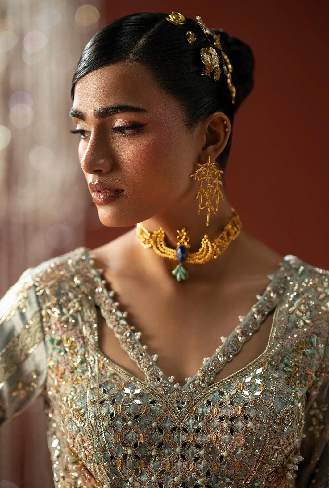 A woman wearing ornate gold jewelry, including a choker necklace with a peacock pendant and matching earrings, paired with a heavily embellished silver and gold embroidered traditional blouse.