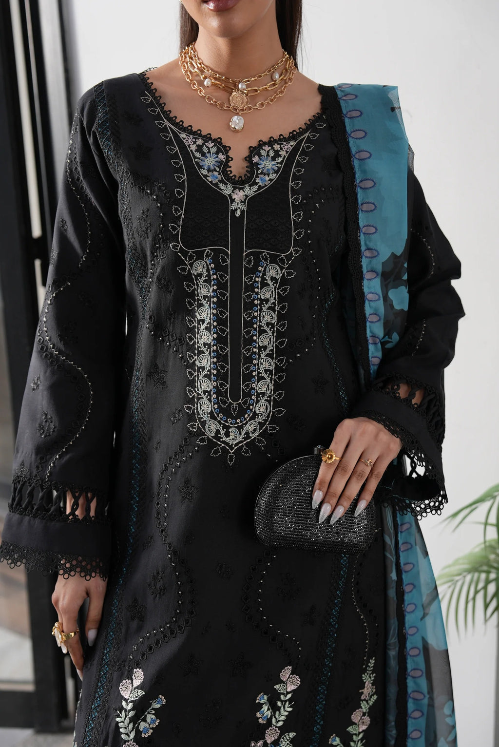 Woman wearing a black embroidered dress with a blue scarf, holding a clutch.
